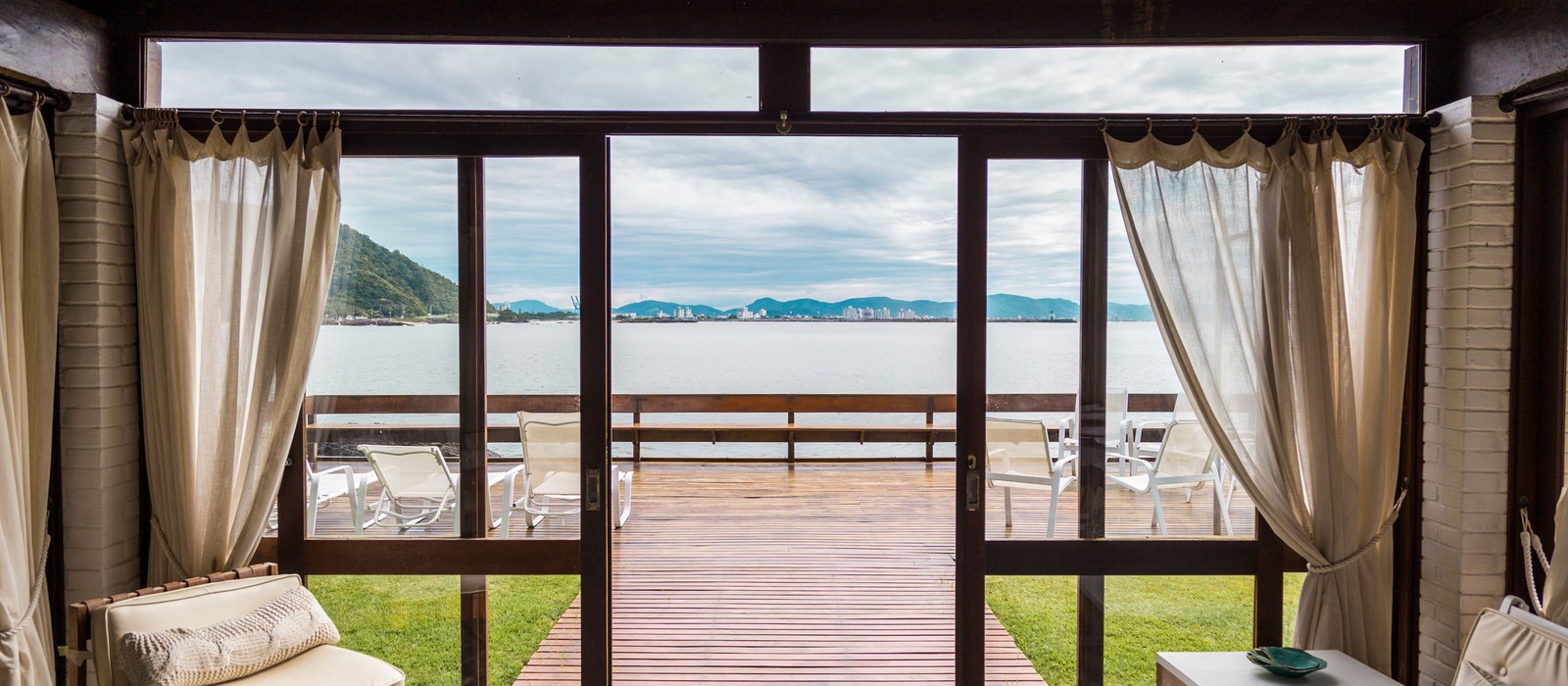 Open glass patio doors with linen curtains drawn back, looking out over a wooden deck to open water and hills