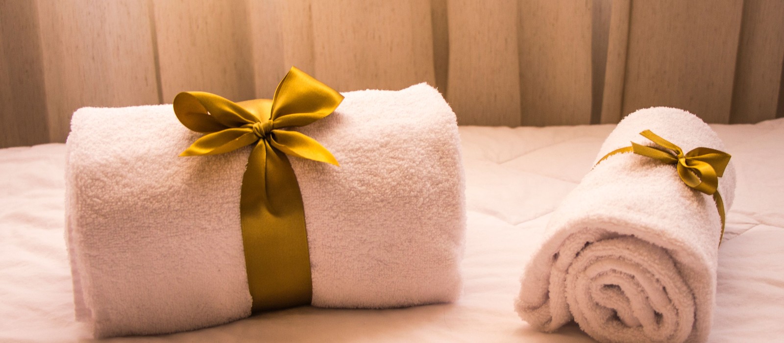 Two rolled white cotton towels tied with gold ribbon, resting on a made bed